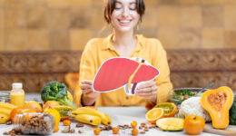 Woman Smiling and Holding a Model of a Human Liver at a Table Full of Healthy Foods Can Fatty Liver Disease Be Reversed